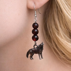 May include: A silver-tone earring with three dark red beads and a howling wolf charm. The earring is suspended from a silver hook and is worn on a person's ear.