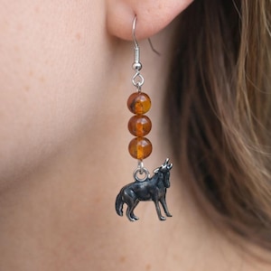 May include: A dangling earring featuring three round, translucent amber-colored beads stacked vertically, suspended from a silver-toned hook. At the base of the beads hangs a small, detailed silver-toned charm shaped like a howling wolf.