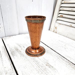 May include: A copper goblet featuring a hammered texture on the main body. The vessel includes a dark metallic band near the rim and a matching base, both accented with small, evenly spaced circular studs around the circumference of the metal.