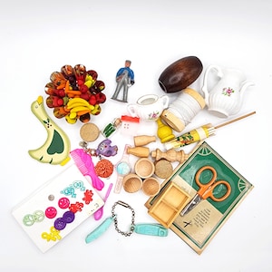 May include: A collection of miniature items including a yellow duck, a fruit bowl, a small figurine, a wooden barrel, a white ceramic pitcher, a spool of white thread, a green book with orange scissors, various wooden bowls, and colorful plastic buttons.
