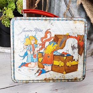 May include: Vintage metal lunchbox with a red handle. The lunchbox features a colorful illustration of two girls and a treasure chest. The words "Junior Miss" are written across the top. The lunchbox has a blue and white striped border.