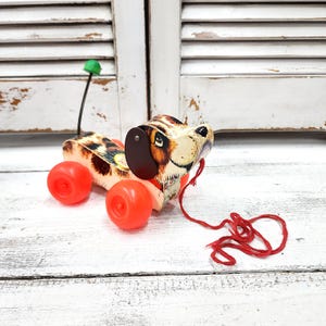 May include: A vintage wooden pull-along toy dog with a spotted body, orange wheels, and a red pull string. The dog's head is painted with a detailed dog face, and it has a green antenna on its back. A classic children's toy.