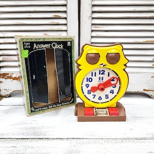 May include: A yellow owl-shaped Answer Clock toy with a white clock face, red hands, and brown accents. The clock has a brown base with red buttons and the text "Answer Clock". The box is green and black.