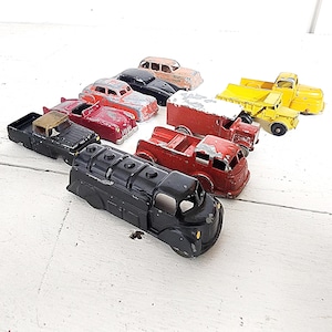 May include: A collection of vintage toy cars and trucks in various colors, including red, black, yellow, and brown. The toys show signs of wear, suggesting they are antique or well-loved items. The collection includes a tanker truck, a pickup truck, and several classic cars.