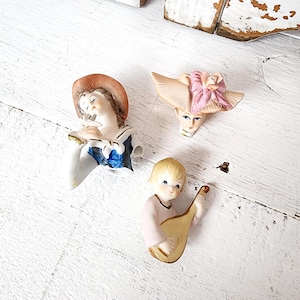 May include: Three vintage porcelain figurines. One depicts a person in a sailor outfit, another a woman wearing a hat with a pink bow, and the third a child playing a lute. The figurines are painted in soft colours.