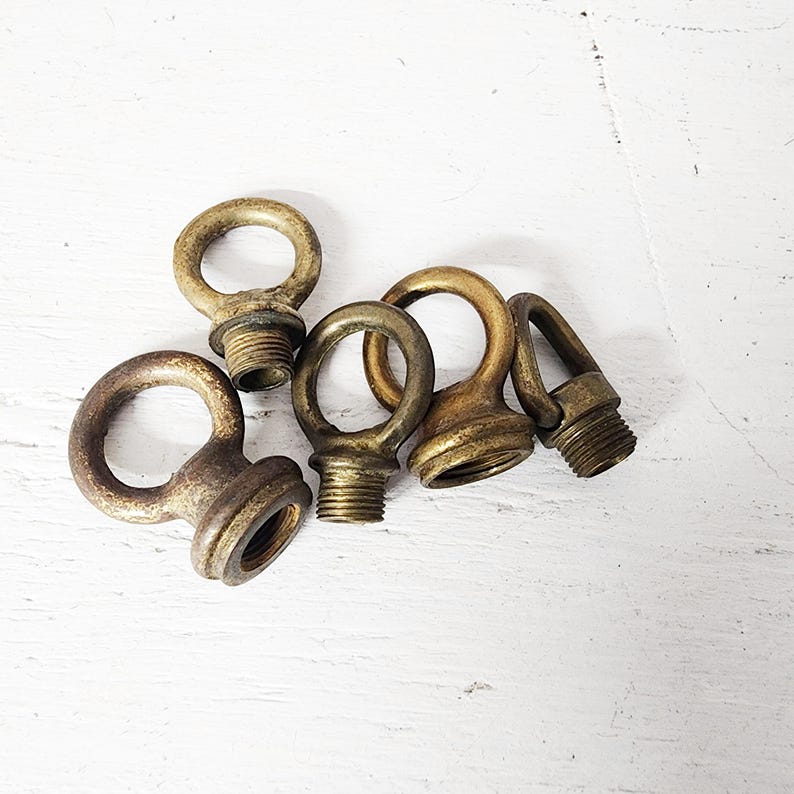 May include: A collection of five antique brass eye bolts with threaded bases. The eye bolts are in varying shades of gold and bronze, with visible signs of age and wear. They are arranged on a white, textured surface.