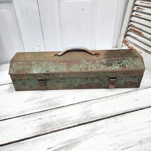 May include: An antique green metal toolbox with a rusty, distressed finish. The rectangular box has a curved metal handle and two clasps. The toolbox is sitting on a white wooden surface.