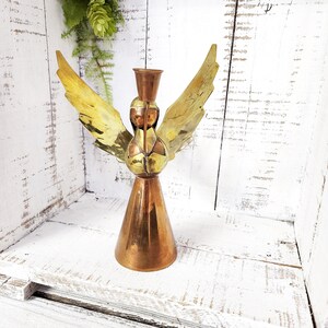 May include: A vintage angel-shaped candle holder. The candle holder is made of copper and brass, with a conical base and outstretched wings. The wings have a gold and brown patina. The angel's head has a small cup to hold a candle.