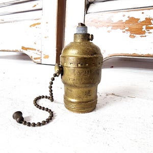 May include: A vintage brass light socket with a pull chain. The socket is marked with the word "EAGLE" and voltage details. The pull chain has a decorative end. The socket is made of brass.