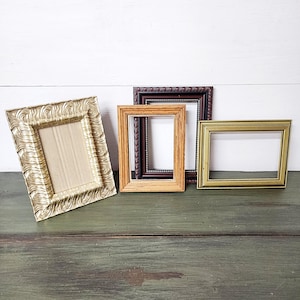May include: Four decorative picture frames in various styles and colors. A gold ornate frame, a brown frame with a beaded edge, a light brown frame, and a gold rectangular frame are displayed on a green surface.