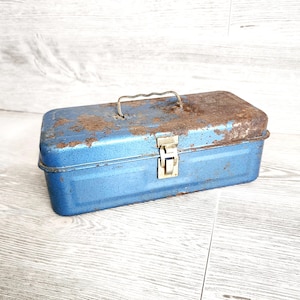 May include: A vintage blue metal toolbox with a silver handle and latch. The top of the toolbox shows signs of rust. The rectangular box is a classic design for storing tools and hardware.