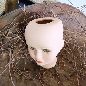 May include: A vintage doll's head with green eyes, long eyelashes, and red lips. The porcelain head is a pale pink colour and has a hole at the top. It rests on dried twigs inside a rusty metal container.