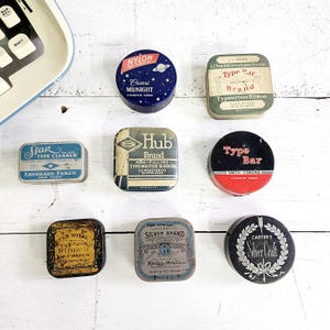 May include: A collection of vintage typewriter ribbon tins and a typewriter keyboard. The tins are various shapes and sizes, with labels featuring brand names like "Nylon," "Hub," and "Silver Brand." The color palette includes blue, black, gold, and green.