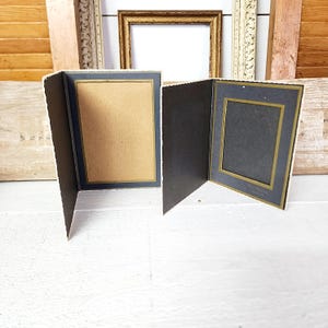 May include: Two vintage photo frames, one open and one closed, with a dark blue border and gold trim. The frames are set against a backdrop of other antique frames and a white wooden surface, creating a nostalgic aesthetic.