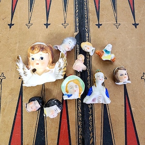 May include: A collection of vintage porcelain doll heads and figurines. The figurines include an angel with gold-tipped wings, a small doll, and several doll heads with painted features. The items are arranged on a brown surface.