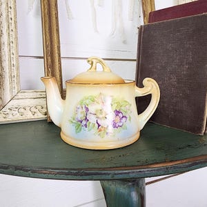 May include: A vintage porcelain teapot with a floral design in shades of white, purple, and green. The teapot has a lid and a handle. It is sitting on a green wooden table.