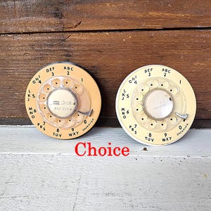 May include: Two vintage rotary phone dials in cream and off-white. Each dial has a circular design with numbers and letters. One dial has the area code 314 and phone number 842-2598. The word "Choice" is written in red.
