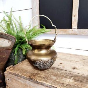 May include: A vintage brass pot with a decorative handle. The pot has a rounded body and a flared rim. A white floral design is etched on the front. The pot is sitting on a wooden crate.