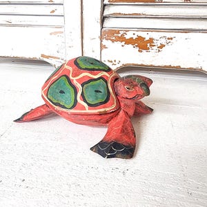 May include: A hand-carved wooden turtle figurine. The turtle has a red shell with green and black accents. The head and flippers are red with black details. The figurine is on a white surface.