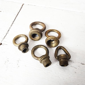 May include: A collection of five vintage brass eye bolts with threaded bases. The eye bolts are in various sizes and show signs of age, with a warm, antique finish. They are arranged on a white wooden surface.