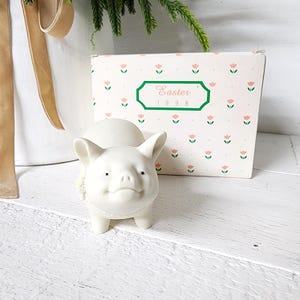 May include: A white ceramic pig figurine with a smiling face and small black eyes. The pig has a decorative collar and stands in front of a box with a floral pattern and the text "Easter 1998".