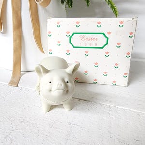 May include: A small, off-white ceramic pig figurine with a textured collar. The pig is smiling and has small black eyes. Behind the pig is a box with a floral pattern and the word "Easter" and the year "1998" printed on it.