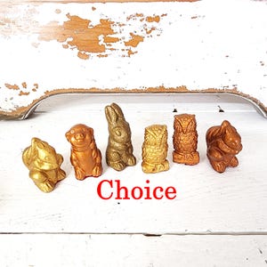 May include: A collection of six small animal figurines in shades of gold and bronze. The figurines include a frog, pig, rabbit, owl, and squirrel. The word "Choice" is written in red text below the figurines.