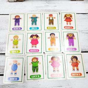 May include: A collection of twelve vintage Cabbage Patch Kids trading cards. Each card features a different doll illustration with names like Burt R., Joe B., and Mary L. The cards have a white border with a green trim and are arranged on a white surface.