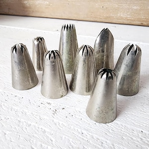 May include: A collection of eight vintage metal piping tips, in various sizes and shapes, for cake decorating. Some tips have the "Wilton" brand name and size numbers like "506" and "2F" etched on them. The tips are silver and sit on a white surface.