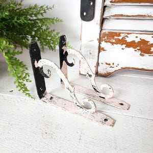 May include: Two distressed, decorative shelf brackets. The brackets are black and white with a pinkish-white base. The brackets have a scrolled design and are made of metal. The brackets are designed to be mounted on a wall.