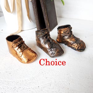 May include: Three bronze-colored baby shoe figurines. The shoes are in different shades of bronze, with laces and detailed stitching. The word "Choice" is written in red. The shoes are on a white surface.