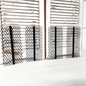 May include: Two black metal diamond-patterned screens with vertical support bars are displayed against a backdrop of distressed white wooden shutters. The screens are positioned on a white surface.