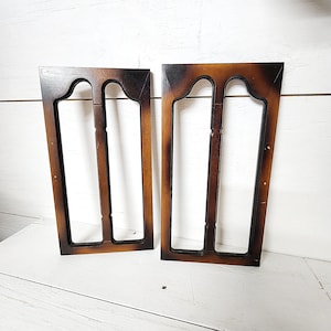 May include: Two wooden decorative frames with a dark brown finish. Each frame has a rectangular shape with an arched top and vertical supports, creating an open, window-like design. The wood grain is visible, adding to the vintage aesthetic.