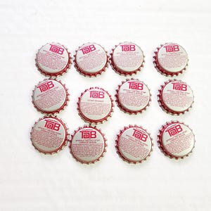 May include: A collection of twelve Tab soda bottle caps. The caps are silver with a red border and feature the Tab logo and text. The caps are arranged on a white surface.