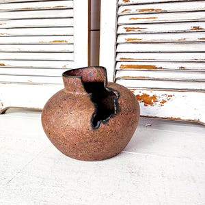 May include: A handcrafted, round ceramic vase with a textured, reddish-brown exterior. The vase has a unique, irregular opening on its side, revealing a dark interior. The rim has a dark, metallic finish.