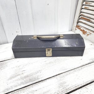 May include: A vintage, dark blue metal toolbox with a brass handle and clasp. The rectangular box has a slightly worn appearance, suggesting age and use. The toolbox is designed for carrying tools and equipment.