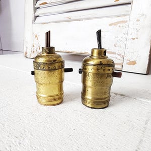 May include: Two vintage brass light bulb sockets with black and brown accents. The sockets have a cylindrical shape and are marked with the brand name "Leviton". The background features a white wooden shutter.