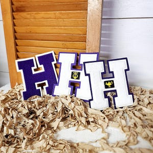 May include: Three letter "H" patches in purple and white. Two of the patches have small embroidered images of a harp and a lamp. The patches are on a bed of brown paper, with a wooden shutter background.