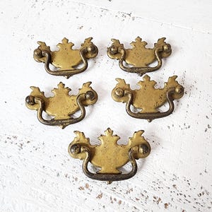 May include: Five antique brass drawer pulls with a decorative, ornate design. Each pull features a curved handle and a central, stylized floral element. The brass has a warm, aged patina, suggesting vintage charm. Perfect for furniture restoration projects.