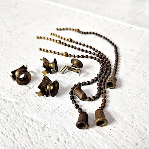 May include: A collection of vintage brass lamp parts, including pull chains with bell-shaped pulls, decorative brackets, and mounting hardware. The components are arranged on a white, textured surface, showcasing their aged patina.