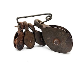 May include: A collection of four graduated dark brown metal pulleys suspended from a large, bent metal safety pin. Each pulley features a circular wheel encased in a weathered, oxidised metal housing, showing signs of age and heavy industrial use.