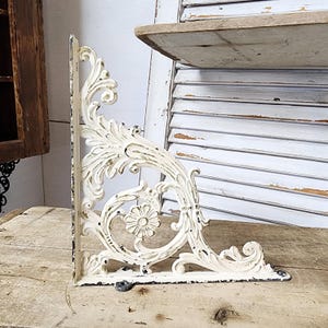 May include: Off-white, ornate metal shelf bracket with a distressed finish. The bracket features a floral and scroll design, with a flat base and a right-angle support. The bracket is designed to support a shelf and has a vintage, antique aesthetic.