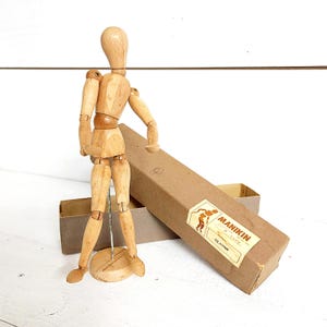 May include: A wooden artist's manikin with articulated joints stands on a round base. The manikin is light brown and is posed with one arm raised. A cardboard box with the word "MANIKIN" printed on it is behind the manikin.