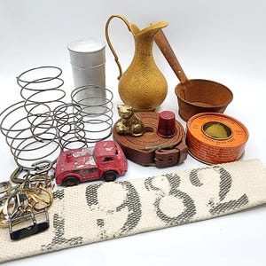 May include: A collection of vintage items including metal springs, a silver canister, a gold-toned etched pitcher, a rusty ladle, a red toy truck, a brown leather belt, a brass cat figurine, a red thimble, thread seal tape, and a white fabric belt with 1982.