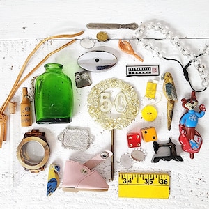 May include: A collection of vintage items including a green glass bottle, a miniature bottle, a watch, a baby shoe, a parrot figurine, a "50" cake topper, dice, and a "Instamatic 104" camera label. The items are arranged on a white surface.