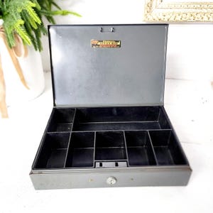Peut inclure: Coffre-fort en métal gris ouvert, avec un intérieur noir divisé en compartiments. Le couvercle est ouvert, révélant l'organisation intérieure. Une petite étiquette est visible sur le couvercle. La boîte a une serrure à clé à l'avant.