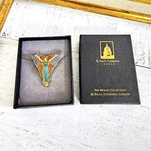 May include: A colorful angel pin with blue, orange, and green enamel, displayed in a black box. The box is next to a black case with gold text that reads "St Paul's Cathedral London" and "The Mosaic Collection".