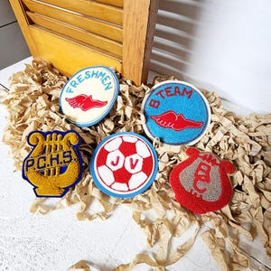 May include: A collection of vintage embroidered patches in various shapes and colors. Includes patches with text "FRESHMEN", "B TEAM", "P.C.H.S.", a soccer ball design, and a red patch with a musical instrument.