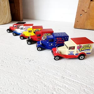 May include: A collection of vintage toy trucks in various colors, including red, blue, yellow, and white. Each truck features circus-themed graphics and logos. The trucks are arranged in a row, showcasing their classic design and detailing.