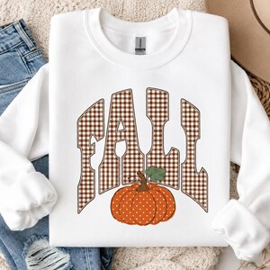 May include: A white crewneck sweatshirt featuring the word FALL in a brown and white gingham check pattern. Below the text is a round orange pumpkin with white polka dots and a green stem and leaf, centred on the front of the garment.